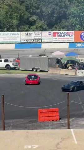 The video shows a car drifting on a race track. The car is red and is being driven around a series of orange cones. There are spectators in the background, as well as other cars and trailers. The race track is asphalt and appears to be outdoors, with trees and a blue and white wall in the background. The video is shot from a slightly elevated position, looking down on the race track.