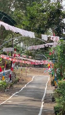 The video shows a paved pathway leading through an archway decorated with white and red and white fabric banners. The pathway is lined with plants and colorful decorations. In the background, a train is visible passing behind the archway. The overall scene appears to be outdoors, with trees and foliage surrounding the pathway.
