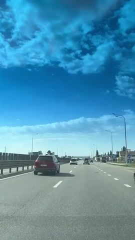 A car drives on a multilane highway under a bright blue sky dotted with fluffy white clouds. Several other cars are also traveling on the highway, going in the same direction. Streetlights line the side of the road. Guardrails are visible on the left side of the road. A billboard and trees are further to the left. Buildings can be seen on the right side of the road. The sky is reflected in the car's windshield, creating a slightly blue tint to the view.