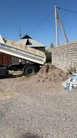 A dump truck is unloading sand into a pile next to a brick wall on a sunny day. The truck's bed is raised, and sand is cascading down into the pile. Two people are standing near the truck. The environment appears to be a construction site or an area where building materials are being delivered.