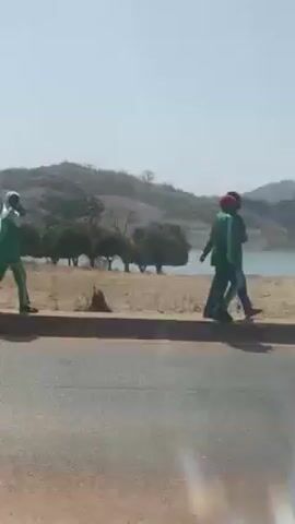 The video shows a landscape view from a moving vehicle. Two people walk along the roadside in the initial frames. Beyond the road, there is a body of water, potentially a lake, with trees along its bank. There are some hills visible in the background. The day is bright, with a clear sky. As the vehicle moves, utility poles become visible along the side of the road.