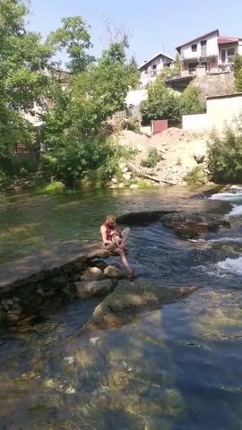 The video shows a woman sitting on rocks in a shallow river with a baby in her lap. The woman is wearing a maroon top and patterned shorts. The baby is wearing a diaper. The river has clear water and visible rocks. Behind the river, there are trees, dirt, and houses on a hillside. The sky is blue and the sun is shining. The camera is positioned at eye level, looking towards the woman and baby.