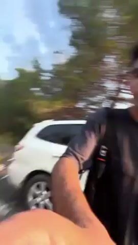 Here's a description of the video:

The video appears to be a selfie taken by a young man outdoors. He has short dark hair, wears glasses, a dark t-shirt, and a black vest. He's standing next to a white car parked on the side of a road. 

In the background, there are other vehicles parked or moving on the road. There also appears to be smoke visible in the distance, partially obscuring the sky and suggesting something is burning nearby. The environment appears to be somewhat rural, with trees lining the road. The sky is bright, suggesting it's either early morning or late afternoon.