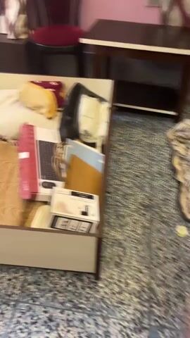 The video shows an unfurnished room with light blue speckled flooring. A dark brown wooden bed frame is in the center of the room. A portion of the bed frame is assembled, with the side rails and support beams visible. Another part of the bed frame, which appears to be a storage drawer or headboard, is partially assembled and lies on its side. The room also contains a partially visible piece of furniture with a dark brown exterior and a lighter interior, possibly a chest of drawers or nightstand. The walls are painted in a pale pink hue. The lighting in the room is uneven, with some areas appearing brighter than others. The camera is handheld and moves erratically, giving a shaky, unpolished feel to the footage. The video seems to capture the process of assembling or disassembling the bed frame.
