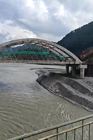 A partially constructed arched bridge spans a muddy river under a cloudy sky. The bridge has a metal support structure forming an arch, with scaffolding and green netting visible in the center. One side of the bridge rests on concrete supports that are partially submerged in the water, with some erosion visible around them. The other side appears to be incomplete, with missing sections of roadbed and supports. The river is wide and appears to have a strong current, with debris and sediment visible along the banks. The surrounding landscape is mountainous and covered in vegetation. The video pans across the bridge and river.