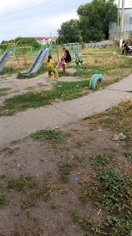 Two children are in a park with playground equipment. A child in a yellow outfit is standing near a seesaw. Another child is sitting on the seesaw. There are two slides in the background. A motorcycle is visible in the far right background.