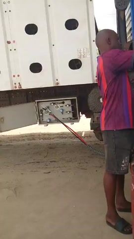 Here's a description of the video:

The video shows a series of shots depicting people fueling a large truck with gas at what appears to be a gas station.

Shot 1 focuses on a man in a purple and red shirt connecting a hose to the truck's fuel system. The truck is white and blue.

Shot 2 shows two men observing the connection. One man is standing while the other is seated. The blue truck's wheel is visible in the background.

Shot 3 shifts to a gas pump with a stainless steel finish. The unit appears worn with peeling labels.

Shot 4 shows a woman sitting on a stool near the gas pump, seemingly working on something in her hands. Hoses are connected to the pump. Other people are sitting in the background.

The environment suggests an outdoor filling station with a paved area.
