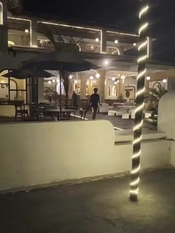 The video shows the exterior of a building at night. The building appears to be a restaurant or cafe with outdoor seating. There are tables and chairs set up outside, some with umbrellas for shade. The building is illuminated with string lights, and there is a prominent pole wrapped in string lights in the foreground. Two people are visible walking in the distance. The overall atmosphere appears to be calm and inviting.