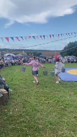 A group of people are participating in an outdoor event on a grassy field. Three young women are at the center of the frame, walking away from the camera. Each woman is carrying a bucket. They appear to be competing in a race or game. The woman on the left is wearing a pink shirt and denim shorts. The woman in the middle is wearing a floral dress. The woman on the right is wearing a white t-shirt and shorts. They are walking on a mowed lawn. In the background, there are many spectators sitting in chairs and on the ground, watching the event. There are tents and stalls set up behind the spectators. Red, white, and blue pennant banners are strung across the sky. A blue and yellow mat is visible on the right side of the frame. The sky is partly cloudy.