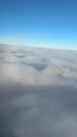 The video shows a view from above a layer of clouds, with a clear blue sky visible above. The cloud cover appears thick and dense, creating a textured landscape that stretches across the horizon. The lighting is bright, indicating daylight, and the overall scene conveys a sense of tranquility and height.