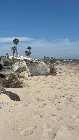 The video pans across a sandy beach. In the foreground, there are numerous footprints in the sand. To the left, large rocks are piled up, with palm trees visible behind them. In the middle ground, the beach extends towards the ocean, where waves are breaking. Further in the distance, a coastline with buildings and more palm trees is visible under a clear blue sky with scattered clouds. The overall scene depicts a sunny day at the beach.