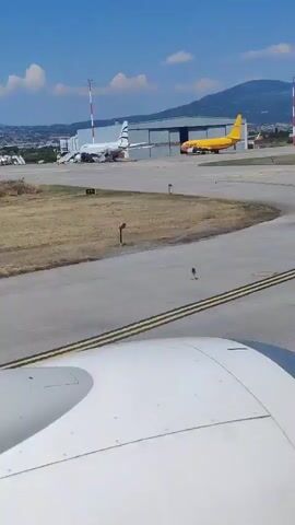 The video shows an airport scene on a clear, sunny day with a bright blue sky and scattered clouds. The viewpoint is from inside an airplane, looking out past the wing. On the tarmac, several aircraft are visible. In the background, a large yellow DHL cargo plane is parked near a hangar. Further in the distance, other smaller planes can be seen on the runway or taxiways. Mountains form a backdrop to the airport, with a town or city visible on their slopes. Red and white striped poles, likely markers or navigation aids, are scattered across the airfield. The time of day appears to be daytime, likely late morning or early afternoon, given the strong sunlight and shadows. The overall atmosphere is one of activity at an airport. The visible text on the tarmac reads "MAX SPE" and "M". The location is confirmed as Neo Rysio, Greece, which is near Thessaloniki Airport (SKG).