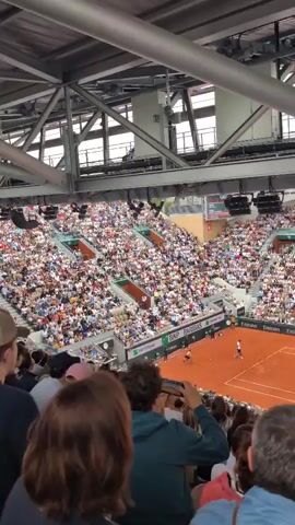 The video shows an elevated view of a tennis match, likely at a major tournament like the French Open given the clay court and the dense crowd. The stadium is packed with spectators, mostly dressed in casual clothing. The camera pans across the court, where two players are visible, although their faces are difficult to discern due to the distance. The court is surrounded by sponsor banners like Lacoste, Renault, and Emirates. The video appears to be taken from a higher seating section, looking down on the match. A portion of the stadium's metal framework is visible overhead. People in the immediate foreground are taking photos and videos of the event. Loud shouts are heard from the crowd.
