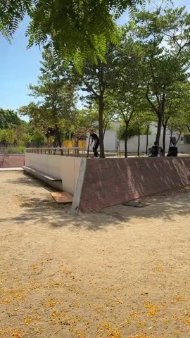 A person on a skateboard rides along a metal rail on top of a concrete wall. The skateboarder wears a black t-shirt and black pants. The skateboarder attempts to do a trick, but fails and falls off the rail. The skateboarder is in a park-like setting with trees and sand.