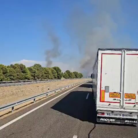 A white truck with red trim on the back is driving on a highway. To the left of the highway is a row of green trees and a dusty embankment. Beyond the trees, a plume of dark smoke is rising into the clear blue sky, indicating a fire. The truck is moving away from the camera, and the smoke is visible in the distance. The license plate of the truck is partially visible.