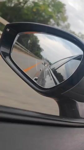 A close-up shot is captured from inside a car, looking at the passenger-side mirror. The mirror reflects a road with a yellow line marking. Trees and a cloudy sky are visible in the reflection, as well as another vehicle. The car appears to be in motion on a highway.