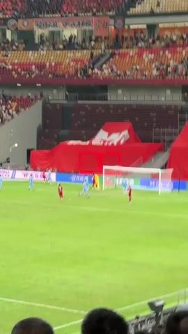 The video shows a soccer game taking place in a stadium with a large crowd. Players in red and light blue jerseys are actively playing on a green field. A goalpost stands at one end of the field, partially obscured by a red tarp. The stadium is filled with spectators, many wearing red clothing and waving red flags. Banners with Chinese characters are displayed in the stands. The camera pans across the scene, focusing on the game and then shifting to capture the enthusiastic crowd.