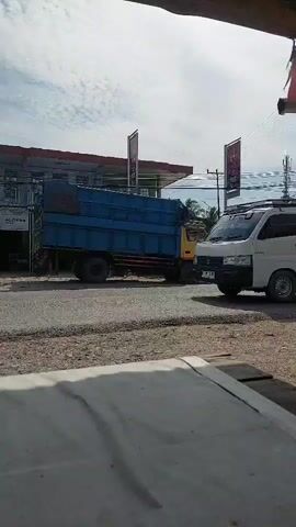 A blue and yellow truck is parked on the side of a road. The truck is carrying a load of green and blue objects. The truck is parked in front of a building that appears to be a gas station. There are power lines and trees in the background. The sky is cloudy.