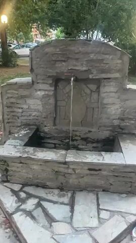 A video shows a stone fountain with water running from a faucet. The fountain is made of rough-hewn stones and has a carved decorative panel in the background. Water flows from the faucet into a basin below. The area around the fountain is paved with flagstones. Trees and buildings are visible in the background. The video is taken from a slightly low angle, looking up at the fountain. The lighting suggests it is daytime.