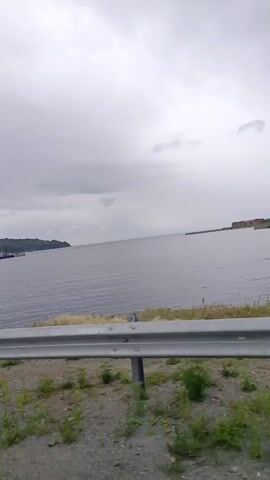 The video shows a view from a moving vehicle, looking out over a body of water. In the distance, along the shoreline, there are several boats docked and a tree-covered landmass. The sky is cloudy, and the water appears calm. A metal guardrail is visible in the foreground, and the ground consists of gravel and sparse vegetation.