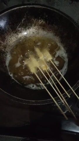The video shows a close-up of food being fried in oil. Initially, the camera focuses on a dark, round pan with bubbling hot oil. Several skewers with food items, possibly spring rolls or fried dough, are submerged in the oil. The food appears to be golden brown and is actively cooking. The camera then pans slightly to reveal the skewers being lifted out of the pan and placed on a metal strainer with holes. The strainer is positioned over what appears to be a countertop or sink area. The food items on the skewers are a uniform golden-yellow color, suggesting they are fully cooked and ready to be drained. The background is dark and somewhat out of focus, with hints of colorful objects, likely kitchenware, visible. The lighting is dim, suggesting the cooking is taking place in an indoor setting, possibly at night. The overall impression is a home-cooking or small-scale food preparation scene.