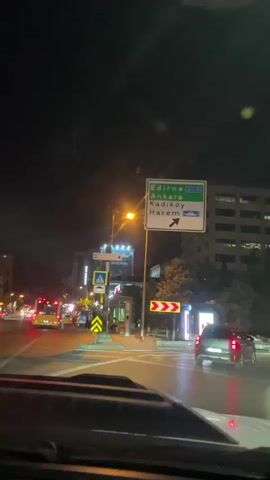 The video is a first-person perspective from inside a vehicle at night, looking forward and slightly to the right. The vehicle is stopped at a traffic light or intersection. A large green directional sign is visible, indicating directions to Edirne, Ankara, Kadıköy, and Harem. Streetlights illuminate the scene, and the road appears wet, suggesting recent rain. There are other vehicles on the road, including cars and a bus in the distance. Buildings are visible in the background, some with illuminated windows. Trees and traffic signals are also present. The overall atmosphere is urban and nocturnal.