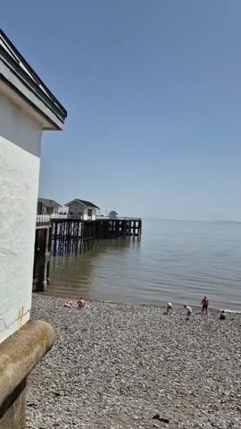 The video shows a pebble beach on a sunny day. In the distance, a pier extends into the calm water. Several people are scattered along the beach, some near the water's edge and others further up the beach. Two individuals are bending down, seemingly occupied with something on the ground.  The camera pans to the right, revealing more of the beach and an island in the distance. More people are visible, some walking along the beach. The camera's viewpoint is slightly elevated.