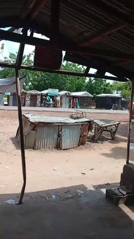 The video pans across a dusty market scene. In the foreground, a makeshift stall with a corrugated metal roof and sides is visible. A wooden table with a crate on it stands to the right of this stall. Beyond these, several other stalls with corrugated metal roofs are lined up. Some of these stalls have goods displayed, and a few people are standing or walking around them. In the background, there are trees and more buildings. A motorcycle with two riders passes by in the distance. The overall environment appears to be outdoors, with a dirt or gravel ground. The weather appears to be daytime and possibly warm, given the open-air market setting and sparse clothing on the people.