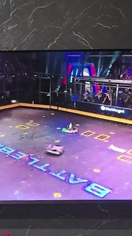 The video shows a robot combat competition. Two robots are in an arena with a purple floor. One robot is predominantly black and silver, and the other is green with pink accents. Sparks are flying from the robots as they engage in battle. In the background, spectators can be seen watching from behind glass enclosures. Two men, one with a beard and one with short blond hair, are visible in the foreground, looking at the arena and appearing to be operating controllers. The arena has yellow markings and blue neon lights outlining the word "BATTLE". One robot is flipped onto its side, while the other is spinning. The scene is chaotic and energetic, indicative of a fight.