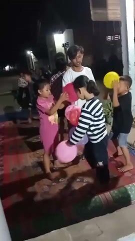 A group of children are standing on a rug outdoors at night. Several of the children are holding balloons. One child in the foreground is wearing a striped shirt and holding a pink and purple balloon. To their right, another child in a pink outfit is holding a red balloon. Further to the right, a boy in a white t-shirt is watching another boy in a black t-shirt. The boy in the black t-shirt is holding a yellow balloon over his head. Other children and adults are visible in the background. The environment appears to be a residential area with buildings and lights visible.