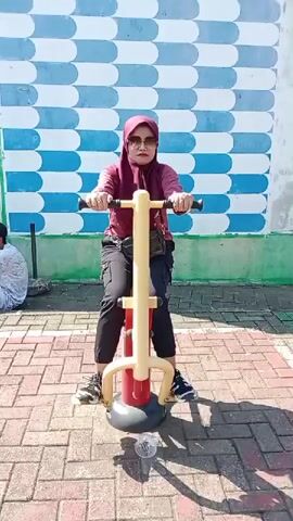 A woman in a maroon hijab and sunglasses is using an outdoor exercise machine. She is wearing a maroon shirt, black pants, and athletic shoes. She is holding onto the handles of the machine and appears to be pumping her legs up and down. The exercise machine is red and yellow with a black base. In the background, there is a wall with a blue and white checkered pattern. To the left of the woman, a person wearing a white shirt and white shorts is sitting on the ground. The ground is paved with brick-like tiles, and there are shadows cast by the sun. A plastic water bottle is visible on the ground near the exercise machine.