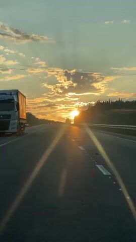 A truck is traveling on a highway during sunset. The sun is low on the horizon, casting long shadows and creating a lens flare effect. The sky is filled with clouds that are illuminated by the sun. Trees and vegetation are visible on either side of the road. The road has lane markings and a guardrail. The image appears to be captured from inside a moving vehicle.