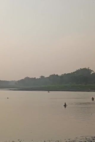 A calm, wide body of water reflects a hazy, pale orange sky. The water's surface is mostly still, with subtle ripples. In the distance, a line of trees with dense foliage forms a horizon. Some of these trees are darker green, suggesting lush vegetation, while others appear lighter or veiled in mist. Several small, dark figures are visible in the water, some appearing to be people fishing. One figure is closer to the foreground, standing in the water, while others are further away. There are also a few small, dark objects scattered on the water, possibly fishing equipment or buoys. The overall atmosphere is serene and natural, with the hazy sky creating a soft, diffused light.
