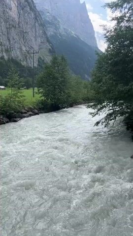 The video shows a wide, fast-flowing river with white, turbulent water. The riverbanks are lined with rocks and trees. In the background, steep, rocky mountains rise towards a cloudy sky. A utility pole is visible on the left bank. A person can be seen briefly in the water on the right side of the frame. The overall impression is a natural, wild landscape.