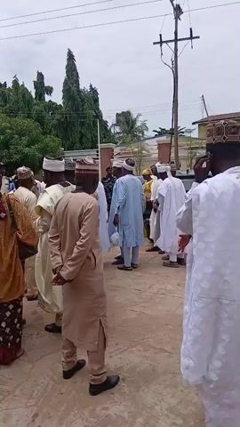 The media file captures a gathering of people in what appears to be an outdoor event or ceremony. The subjects are predominantly men, many of whom are dressed in traditional Islamic attire, including long robes and embroidered caps. Some women are also present, wearing headscarves and colorful patterned dresses. The scene is set in a paved outdoor area, likely a street or courtyard, with buildings and lush green trees visible in the background. A utility pole with electrical wires stands prominently in the frame, suggesting an urban or semi-urban setting.

The activity taking place seems to be a social gathering, possibly a religious ceremony, celebration, or a community event. The people are standing around, some talking to each other, while others appear to be observing something or waiting. Their expressions are difficult to discern clearly due to the distance and resolution, but the overall atmosphere seems calm and orderly.

Notable details include the time of day, which appears to be daytime, judging by the ambient light. The sky is overcast, suggesting cloudy weather. The presence of a green car with a license plate in the foreground provides a modern contrast to the traditional clothing. The license plate displays some text and numbers, but it is not clearly legible. The location context is provided as Sabon Gari North, Nigeria. The people are facing in various directions, indicating a dispersed gathering rather than a directed activity. The overall impression is of a community event taking place in a Nigerian setting.