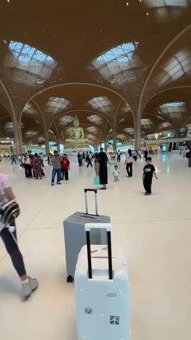 The video begins with a view of a large, open airport terminal. The ceiling is made of a wooden lattice structure with skylights. In the distance, a large golden Buddha statue is visible. Several people are scattered throughout the terminal, some walking and others standing in groups. In the foreground, two suitcases are placed on the polished floor. The camera then pans to the right, showing a woman with long dark hair wearing a black tank top and jeans. She is holding the handle of a suitcase and looking towards the camera. The camera moves with her as she walks, and she smiles. The background shows more of the airport terminal, with large glass windows and more people.