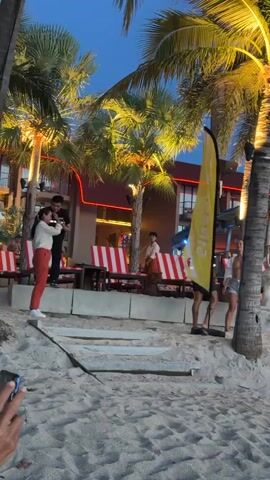 The video shows a beach scene at dusk. A group of people are gathered on the sand, with a building adorned with red and white striped lounge chairs and string lights in the background. Palm trees with illuminated fronds are visible, adding to the tropical ambiance. Some individuals appear to be standing and socializing, while others are seated on the lounge chairs, enjoying the evening. The overall atmosphere suggests a relaxed and lively beachfront gathering.