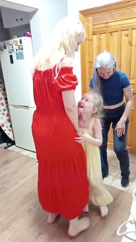 A young girl in a yellow dress hugs the leg of a woman in a red dress. Another woman with grey hair in a blue top stands nearby, looking down. The scene appears to be indoors, possibly a kitchen or dining area, with wooden cabinets and a refrigerator visible.