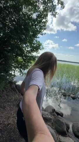 This is a vertical video clip taken at a body of water in Yablonovka, Russia. The scene is set on a sunny day with fluffy white clouds in a bright blue sky. The water is calm, reflecting the sky and surrounding greenery.

The primary subject is a woman with blonde hair, wearing a white t-shirt and dark pants. She is in the foreground, and the camera appears to be held by her or someone very close to her, creating a selfie-like perspective. She turns to face the camera briefly, smiling and looking at the viewer.

The setting is a natural landscape. The foreground consists of the rocky shoreline of the lake, with dry leaves and twigs scattered on the ground. Lush green trees and foliage overhang the water on the left side of the frame, casting shadows. The background shows a vast expanse of water with tall reeds growing along the distant shore. The overall mood is relaxed and pleasant, suggesting a leisurely outdoor activity. The time of day appears to be daytime, likely late morning or early afternoon, given the bright sunlight and clear skies. There is no visible text in the clip.