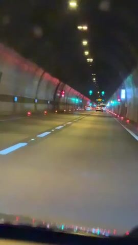 The video shows a view from inside a vehicle driving through a tunnel at night. The tunnel is lit by overhead lights and illuminated by traffic signals. The walls of the tunnel are lined with what appear to be reflective panels or lights. The road ahead is marked with white lane lines, and there are red and green traffic lights visible on both sides of the tunnel. One or two vehicles can be seen in the distance, also driving through the tunnel. The overall impression is of a well-lit and maintained tunnel system.