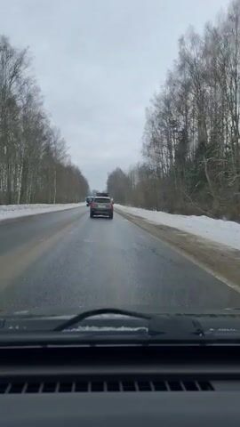 A dashboard camera records a road with two lanes in each direction. The road is wet, with some areas of water reflecting the cloudy sky.  Snow is present on both sides of the road. The road is lined with trees on both sides that do not have leaves.  There are several vehicles on the road ahead of the camera car, including a bus and a gray car. One person is wearing a yellow vest and appears to be on foot.  The interior of the camera car is visible at the bottom of the shot.