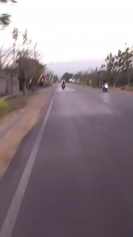 The video shows a first-person perspective of riding a motorcycle down a paved road. The road is lined with trees and some buildings on the left side. There are several colorful flags or banners hanging from poles on the left side of the road. In the distance, a few other vehicles can be seen, including what appears to be another motorcycle and a truck. The sky is overcast and appears to be either dawn or dusk. The video is shaky, suggesting movement.