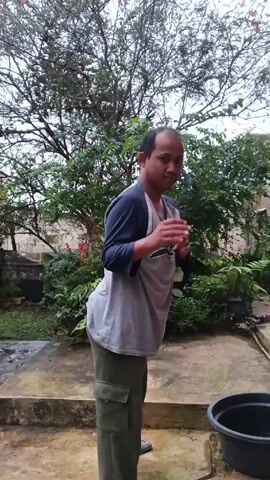 A person stands in a backyard or outdoor area, smiling and making chopping motions with their hands. They have a bald head and are wearing a grey and blue shirt, as well as green pants. The area appears to be a patio or paved space, with plants and greenery in the background. There's also a black plastic tub nearby.