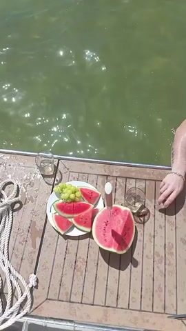 The video begins with a close-up shot of a plate of watermelon slices and grapes on a wooden deck, next to a glass of white wine and a partially eaten watermelon. The water of a lake or ocean is visible in the background. The scene then pans out to reveal a group of people swimming and socializing around a white sailboat anchored in the water. Several individuals are in the water, some standing and some swimming. Others are on the deck of the sailboat, interacting and enjoying the sunny weather. The environment appears to be a natural setting with trees and a shoreline visible in the distance. The overall impression is one of a leisurely summer day spent on the water.