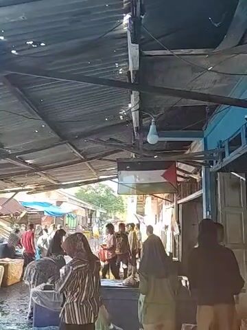 The video shows a bustling outdoor market scene, likely in a tropical or coastal region. The market is covered by a corrugated metal roof structure, which is old and shows signs of wear. A light bulb hangs from the roof. A Palestinian flag hangs prominently, adding a political or solidarity element to the scene. Numerous people are present, walking through the market and interacting with vendors. Some are dressed in casual clothing, while others are wearing traditional or religious attire. A variety of goods are displayed on tables and in containers, possibly including fish, produce, and other food items. The general atmosphere is busy and somewhat chaotic, typical of a lively market. The overall lighting is a combination of natural daylight and artificial lighting.