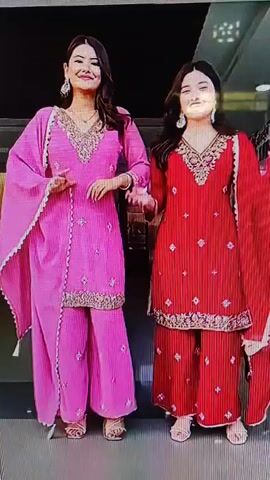 Two women in matching pink and orange floral dresses are dancing. They are smiling and appear to be enjoying themselves. The background is dark, with some blurry lights visible at the top right.