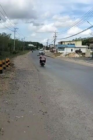 The video shows a paved road with a dirt shoulder on the left. A black motorcycle with one rider is moving away from the camera on the left side of the road. A white SUV is approaching the camera on the right side of the road. There are utility poles with wires lining both sides of the road. Buildings and trees are visible in the background. The sky is cloudy.