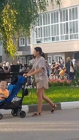 A woman with dark hair pulled back in a bun, wearing a patterned t-shirt and light-colored shorts, pushes a blue and black stroller. She is walking on a paved surface next to a grassy area bordered by a curb. In the background, several people are gathered near a building with large windows. Some individuals are sitting on benches, while others are standing. A few people are near bicycles, and a small group appears to be performing music, with one person playing a guitar and another at a keyboard. The scene is outdoors, with trees and foliage visible. The lighting suggests it is daytime, possibly late afternoon due to the angle of the sun.
