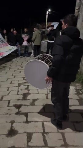 At night, a group of about a dozen people stand on a brick-paved pathway, holding a large white banner with black writing and green and yellow shapes. One person on the right side of the frame holds a large drum and drumstick, preparing to play it. Other people are standing behind the banner and on the periphery of the group, some holding flowers and some recording with their phones. The environment appears to be a residential street with houses in the background. The lighting is dim, primarily from a streetlamp visible in the distance.