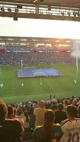 The video shows a packed outdoor soccer stadium from the perspective of spectators in the stands. A large banner, possibly a team flag, is unfurled on the green field, held by many people. Fireworks and smoke are erupting from various points on the field, creating a festive atmosphere. The sun is setting, casting a warm glow over the stadium, and illuminating the sky with orange and yellow hues. The spectators in the foreground are facing the field, some with their backs to the camera, and appear to be engaged in the event. The overall scene depicts a sporting event or ceremony, likely before a game, marked by pyrotechnics and crowd participation.
